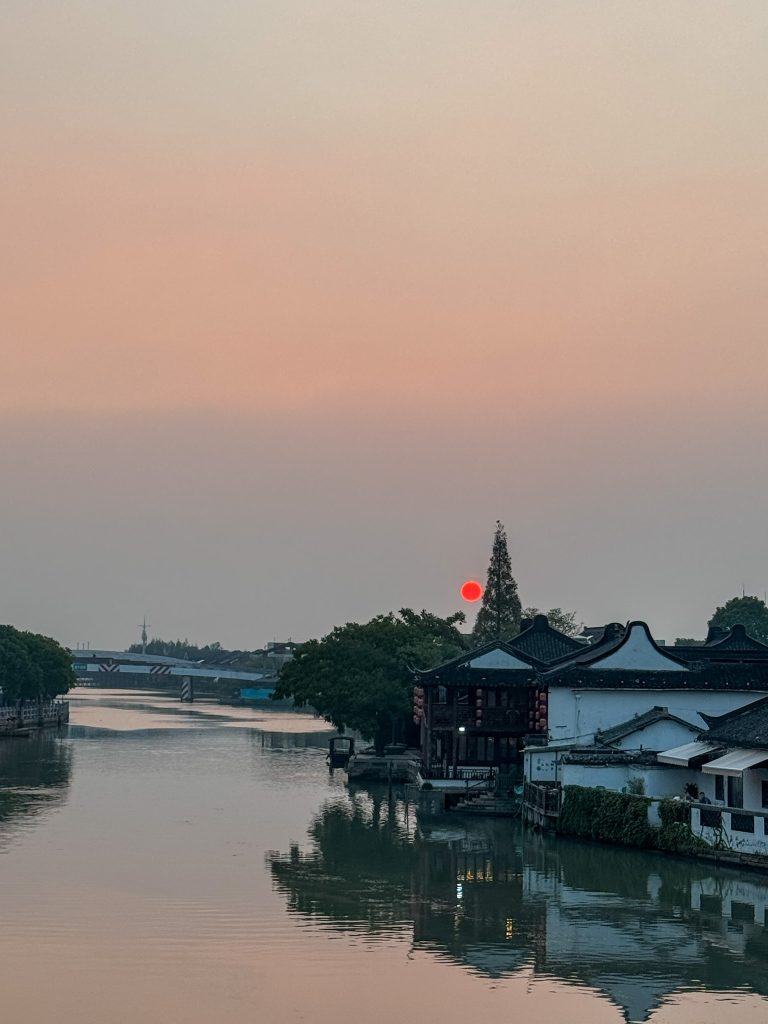 Looking down a water canal in Zhujiajiao Water Town, China at the buildings reflecting in the water and a red moon the in sky