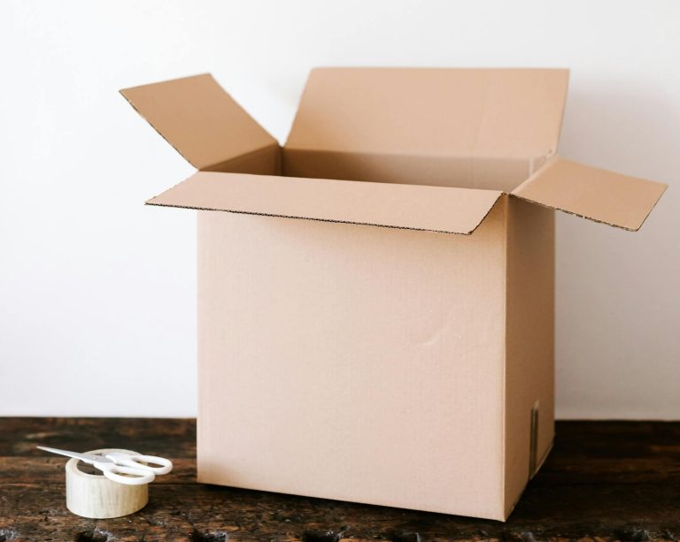 An open cardboard box on a weathered wooden table with tape and scissors beside it, ready for packing.