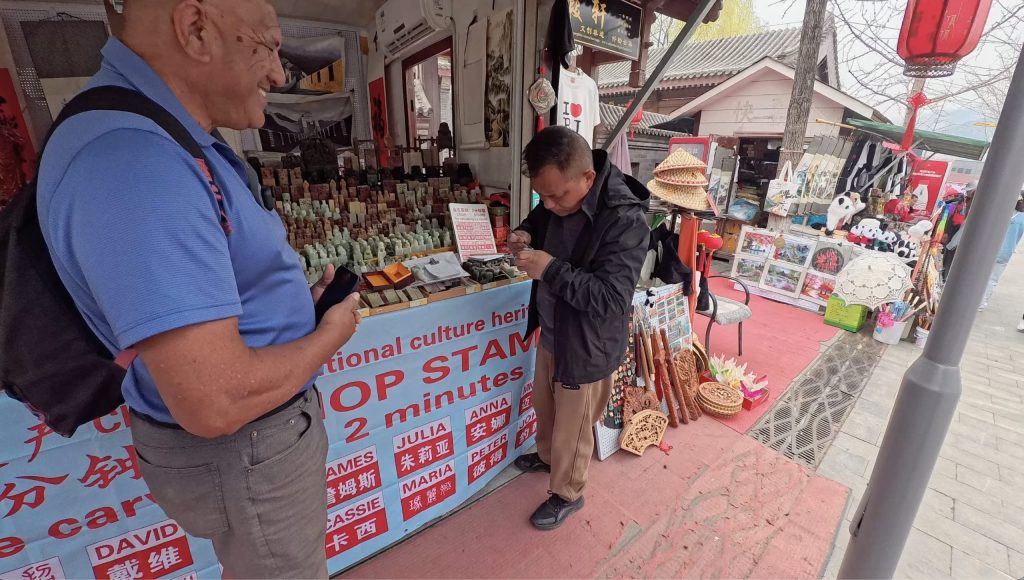 2 men at the shops by the Great Wall of China, one is hand carving a stamp while the other is watching